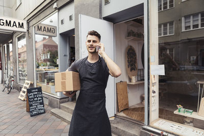 Young male entrepreneur with package box talking on smart phone outside store