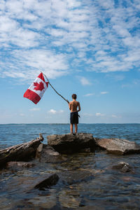 Boy holding canada flag in the air on shore of a lake on a summer day.