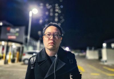 Portrait of young asian man in eyeglasses and earphones walking on city street at night.