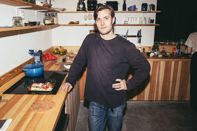 Portrait of man standing with hand on hip while preparing food in kitchen at home