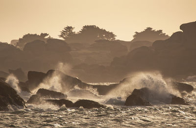 Waves splashing on rocks against sky during sunset