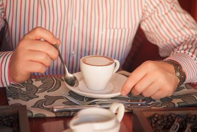 A man in a striped shirt holds a silver teaspoon and a white cup of coffee