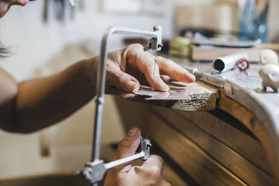 Cropped hands of female artisan using handsaw while making jewelry on wooden table in workshop