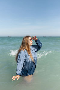 Side view of charming young female in wet shirt standing in sea water and looking into distance in sunny summer day