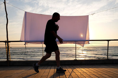 Side view of silhouette man walking on pier at beach during sunset