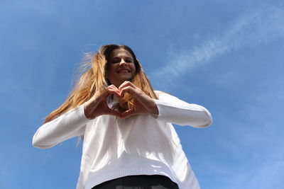 Low angle view of woman making heart shape while standing against blue sky