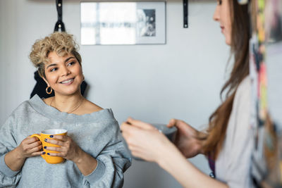 Smiling woman holding mug while looking at friend in room