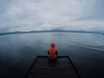 Rear view of person sitting at edge of lake pier against sky