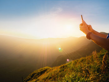 Cropped image hiker of photographing through mobile phone while standing on cliff against mountain ranges during sunset