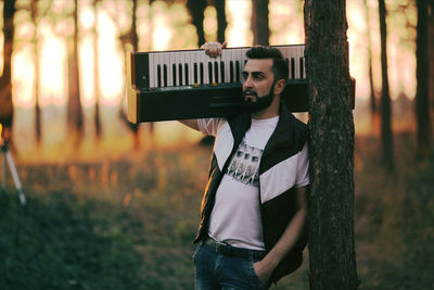 Thoughtful man with keyboard instrument standing by trees in forest