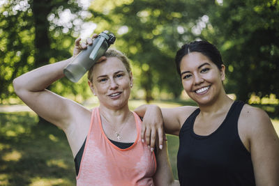 Portrait of smiling woman with exhausted female friend holding water bottle at park