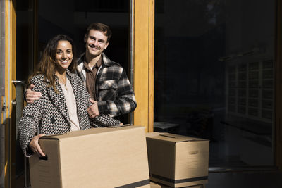 Portrait of smiling man embracing woman holding cardboard box in front of apartment