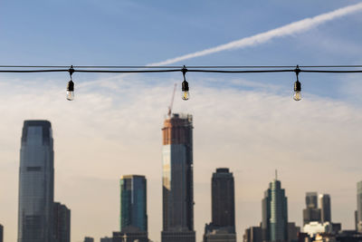 Low angle view of light bulbs hanging against buildings in city against sky