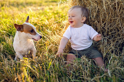 Gay boy kid blonde in white tank top sitting on a field of hay next to stack with dog during sunset