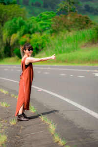 Side view of young woman wearing sunglasses hitchhiking while standing on road
