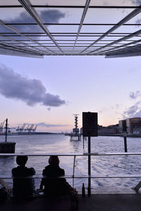 Rear view of people sitting by sea against sky at dusk