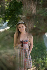 Portrait of smiling young woman wearing hat standing by tree at park