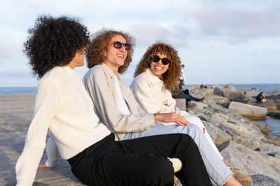 Side view of cheerful friends with curly hair wearing sunglasses sitting close against cityscape and rocky embankment in sunlight