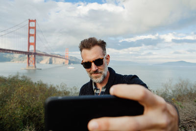 Adult man with beard taking selfie on smartphone with bridge in background