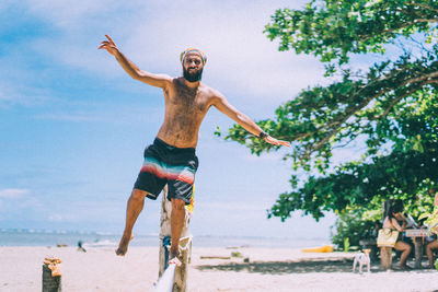 Full length of shirtless man standing on railing at beach against sky