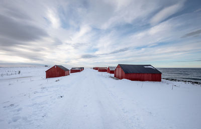 Empty snow covered road amidst barns against cloudy sky