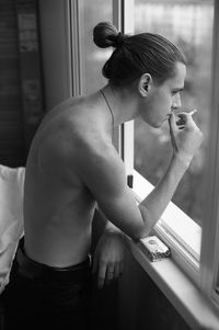 Side view of shirtless young man smoking cigarette by window at home