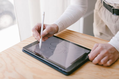 Cropped unrecognizable businesswoman bending over table and working on tablet with stylus in light wooden office