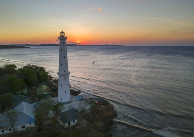 Lighthouse amidst sea and buildings against sky during sunset