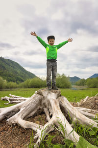 Full length of boy with arms outstretched standing on tree stump against cloudy sky