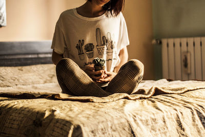 Midsection of woman holding cactus while sitting on bed at home