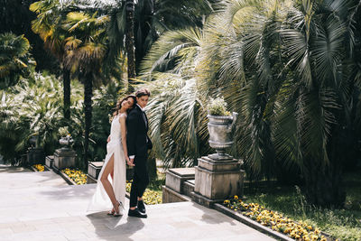 Happy lovers the bride and groom in wedding outfits walk among plants and palm trees in the old park