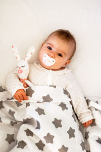 Candid portrait of cute baby girl lying on white bed with pacifier and toy of hare in her hand.