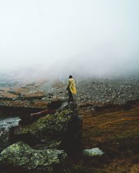 Rear view of man standing on rock against sky during foggy weather