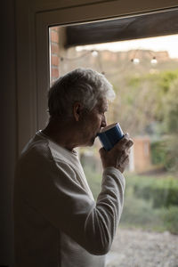 Senior man drinking coffee while standing by window at home