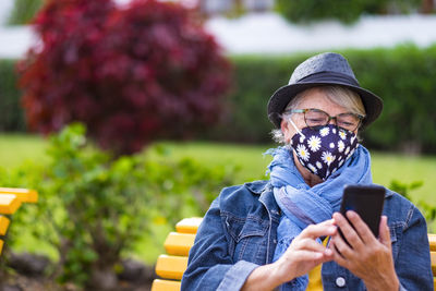 Senor woman wearing mask using mobile phone while sitting outdoors