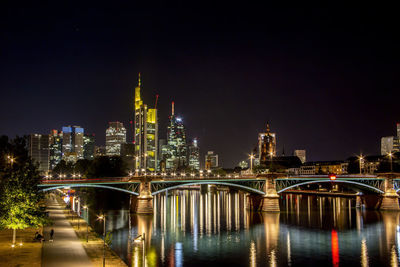 Illuminated bridge over river by buildings against sky at night