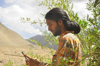 Side view of a young man with long hair using phone with the background of mountains of zanskar