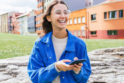 Positive young female in stylish clothes standing in city street and messaging on mobile phone while laughing with closed eyes