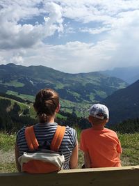 Rear view of mother and son sitting on bench against cloudy sky