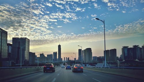 Cars on road by buildings against sky during sunset