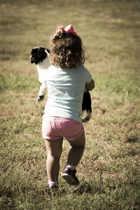 Rear view of girl holding dog while walking on grassy field