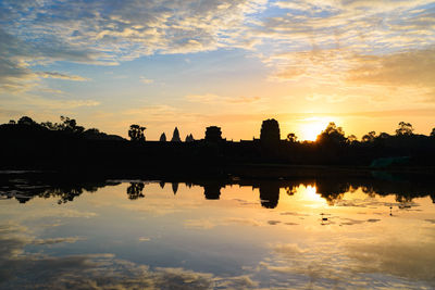 Silhouette buildings by lake against sky during sunset