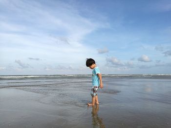 Boy standing on shore at beach against cloudy sky