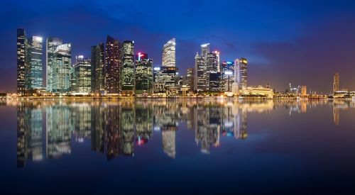 Reflection of illuminated modern buildings on calm river against sky at dusk