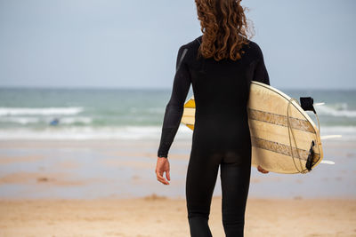 Rear view of woman holding surfboard while walking at beach against sky