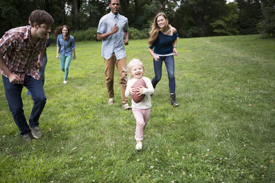 Playful friends running behind girl holding football ball on grassy field