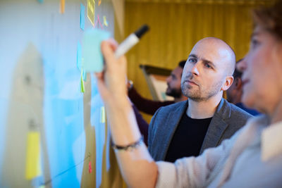 Businessman looking at female coworker removing adhesive note from whiteboard in office seminar