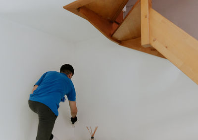 A young oriental man paints the wall under the stairs with a brush.