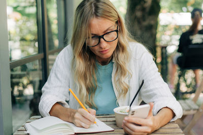 A woman sitting in a cafe at lunch uses a mobile phone and a notepad for work.