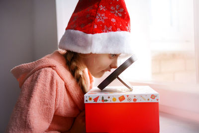 A caucasian girl in a red santa cap looks in the mirror, which stands on a gift, quarantine at home.